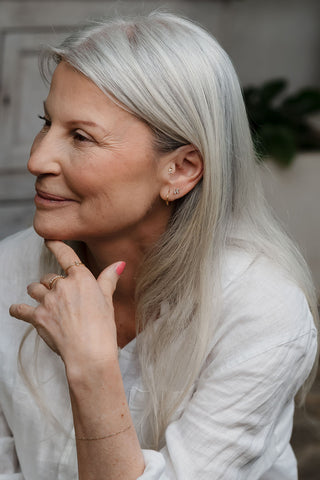 Elegant older woman wearing multiple Tish Lyon gold earrings and rings, styled with a soft white blouse and relaxed smile.